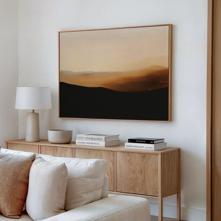 A modern living room features a beige sofa, wooden sideboard with books, a white lamp, and decorative bowls. The Sienna Ridge Canvas Art in warm earth tones hangs above the sideboard, enhancing the contemporary decor.