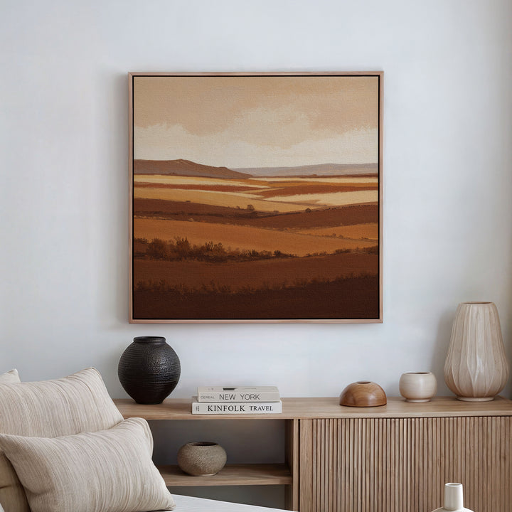 A minimalist living room showcases the Sienna Terrain Canvas Art above a wooden console table adorned with vases, a lamp, and stacked books, while a light-colored sofa is partially visible in the foreground.