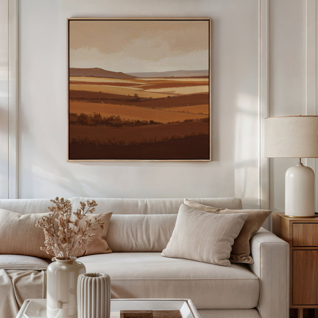 A cozy living room with a beige sofa, neutral cushions, a side table with a ceramic lamp, dried flowers in a vase, and the Sienna Terrain Canvas Art featuring rolling hills in warm brown tones displayed on the wall.