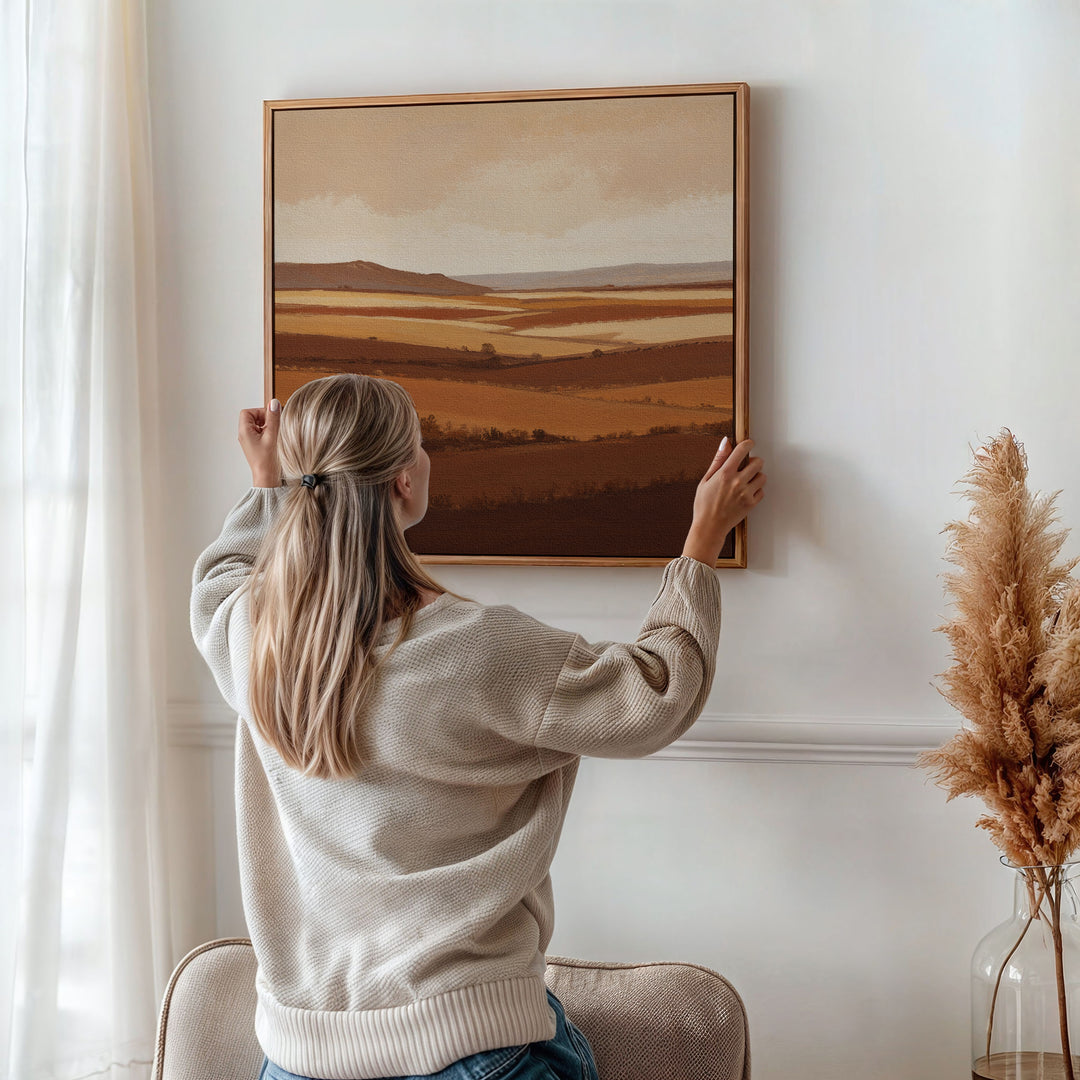 A woman with long blonde hair, dressed in a beige sweater, hangs the Sienna Terrain Canvas Art on a white wall, while dried pampas grass in a vase adds warmth to the softly lit Nordic Park-inspired room.