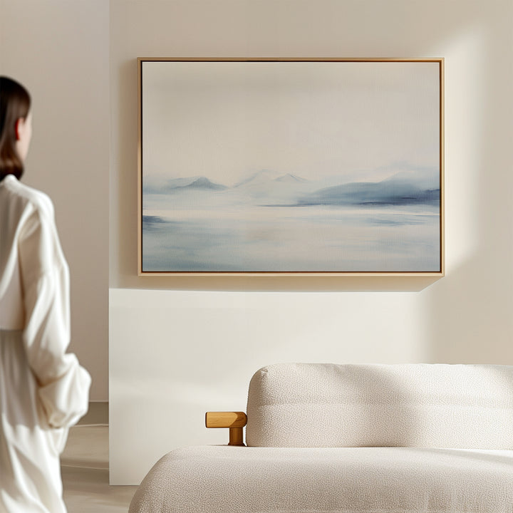 A person stands in a bright, minimal room, admiring Silent Glaciers Canvas Art—minimalist wall art of blue mountains and a misty lake—displayed above a beige sofa.