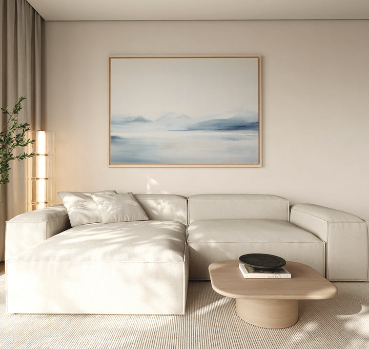 A modern living room with a light beige sectional, low wood coffee table, tall plant, and natural light features Silent Glaciers Canvas Art—large wall art showing blue mountains and water on a light wall.