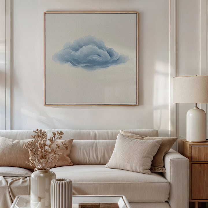 A modern living room with a white sofa, beige pillows, a wooden side table holding a cream lamp and dried flowers, and minimalist wall art featuring the Single Cloud Canvas Art—a large framed painting of a blue cloud.