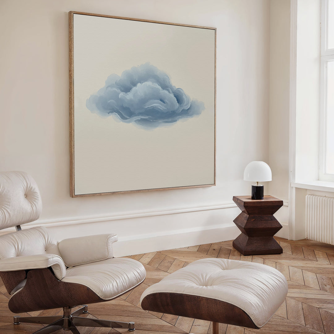 A minimalist living room featuring a white leather chair and ottoman, a small wooden side table with a white lamp, and the Single Cloud Canvas Art on the wall. Light wood floors and cream-colored walls complete the modern decor.