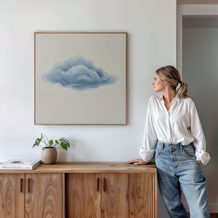A woman in a white blouse and jeans admires the Single Cloud Canvas Art above a wooden cabinet, which holds a potted plant and book, enhancing the modern decor of the minimalist room.