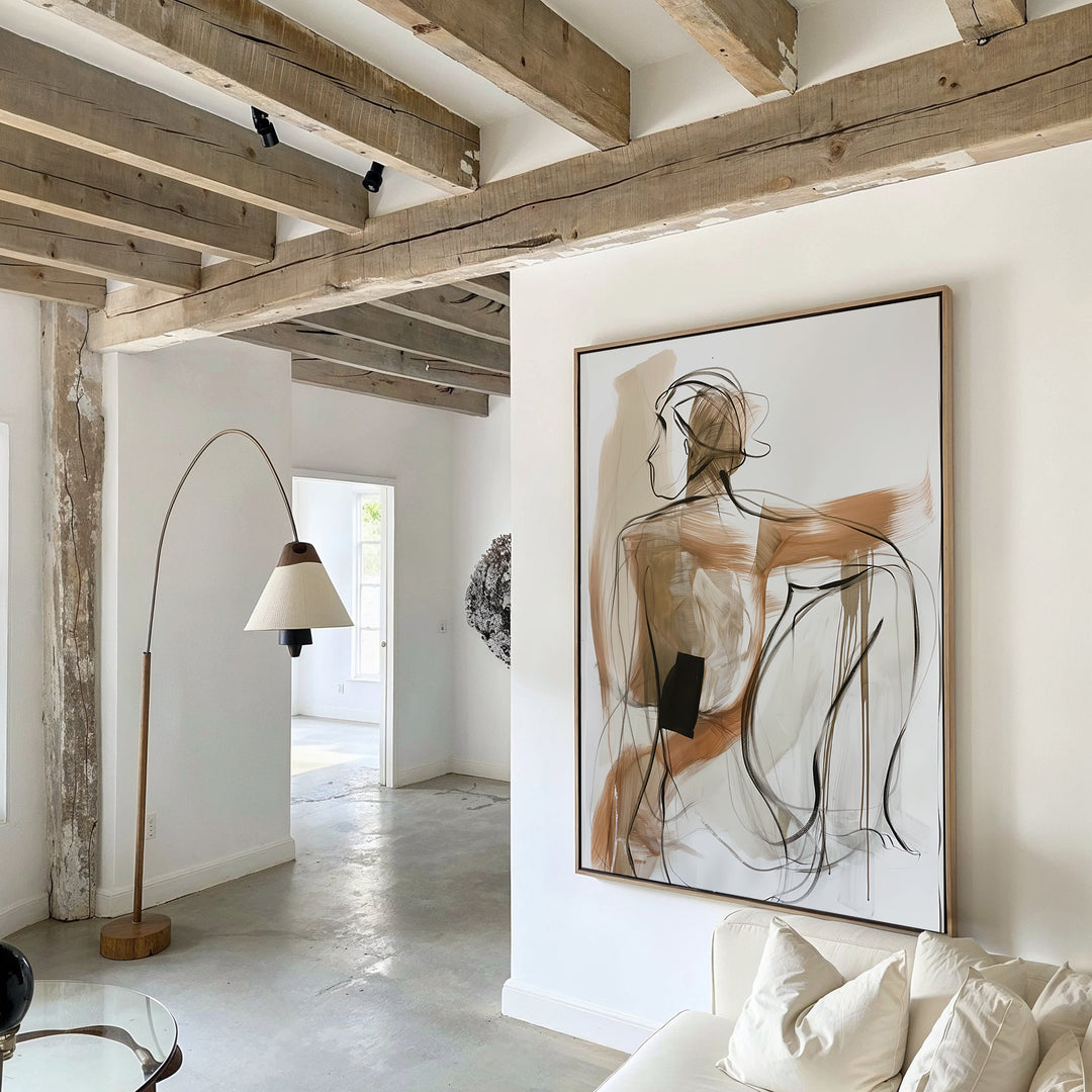 A modern minimalist living room features neutral-toned furniture, exposed wooden beams, and sunlight streaming through an open doorway. The space is highlighted by the Sitting Silhouette Abstract Canvas Art as a striking line art portrait centerpiece.