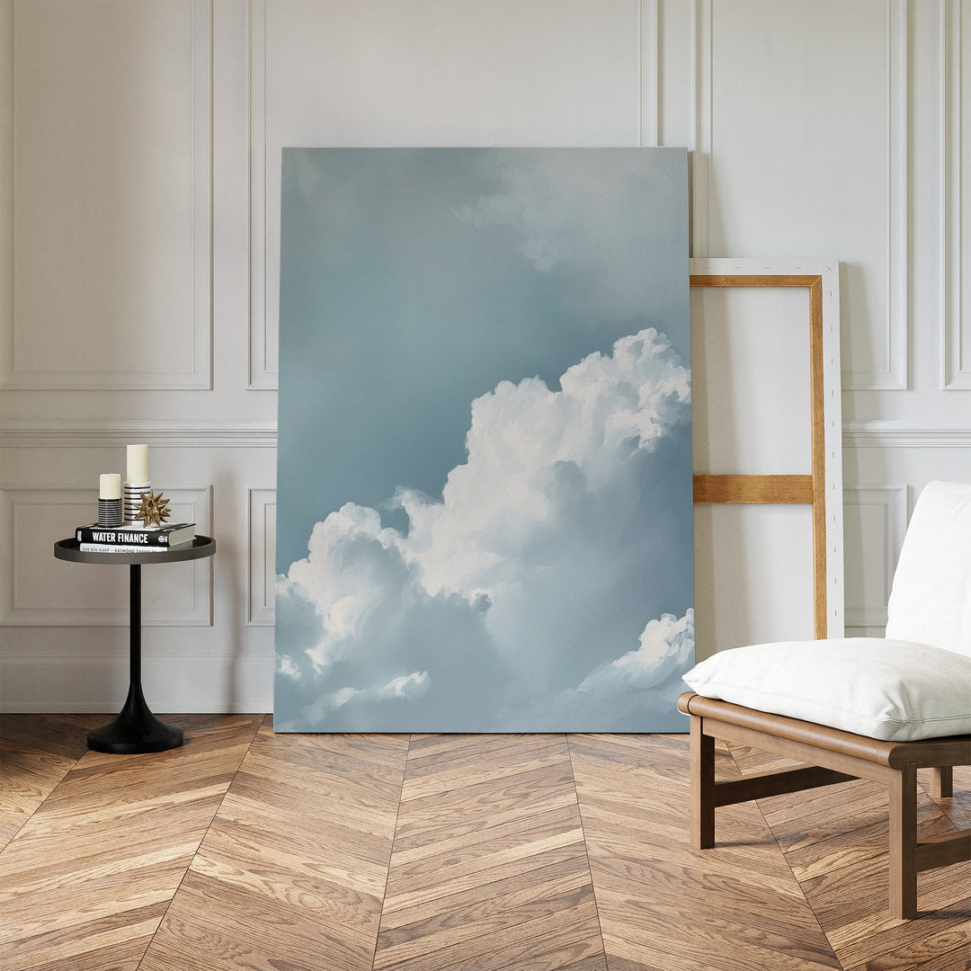 Sky Serenade Abstract Canvas Art, with white clouds in a blue sky, leans against the wall of a minimalist room featuring herringbone wood floors, a modern chair, candles on a table, and an empty wooden frame—ideal for living room decor.