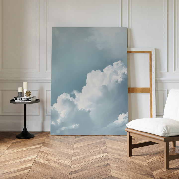 Sky Serenade Abstract Canvas Art, with white clouds in a blue sky, leans against the wall of a minimalist room featuring herringbone wood floors, a modern chair, candles on a table, and an empty wooden frame—ideal for living room decor.