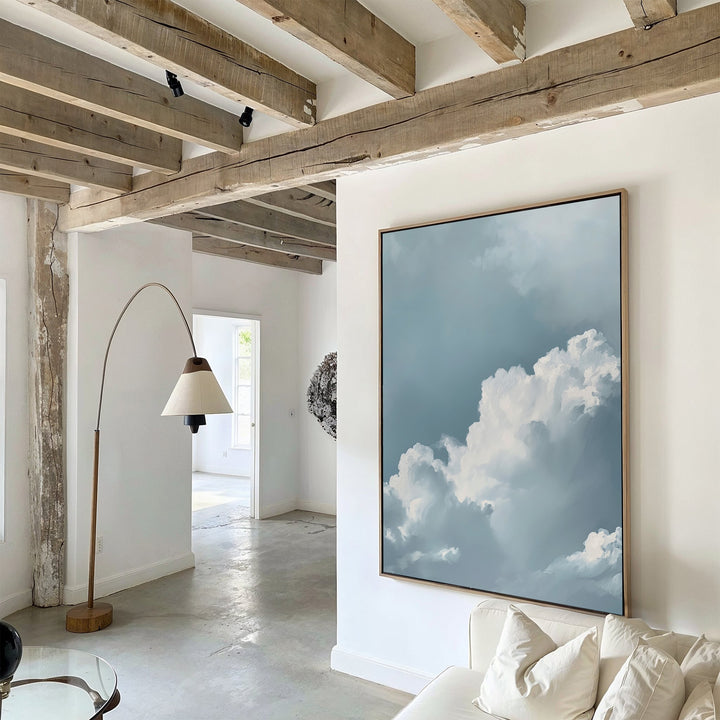 A modern living room with exposed wooden beams, white walls, and Sky Serenade Abstract Canvas Art depicting a blue sky and clouds. A curved floor lamp and white sofa complete the decor, enhanced by natural light from the next room.