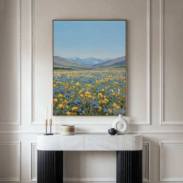 The Skyview Field Canvas Art, featuring a vibrant wildflower field with distant mountains and blue sky, hangs above a modern white and black console table in an elegant paneled room with minimalist decor.