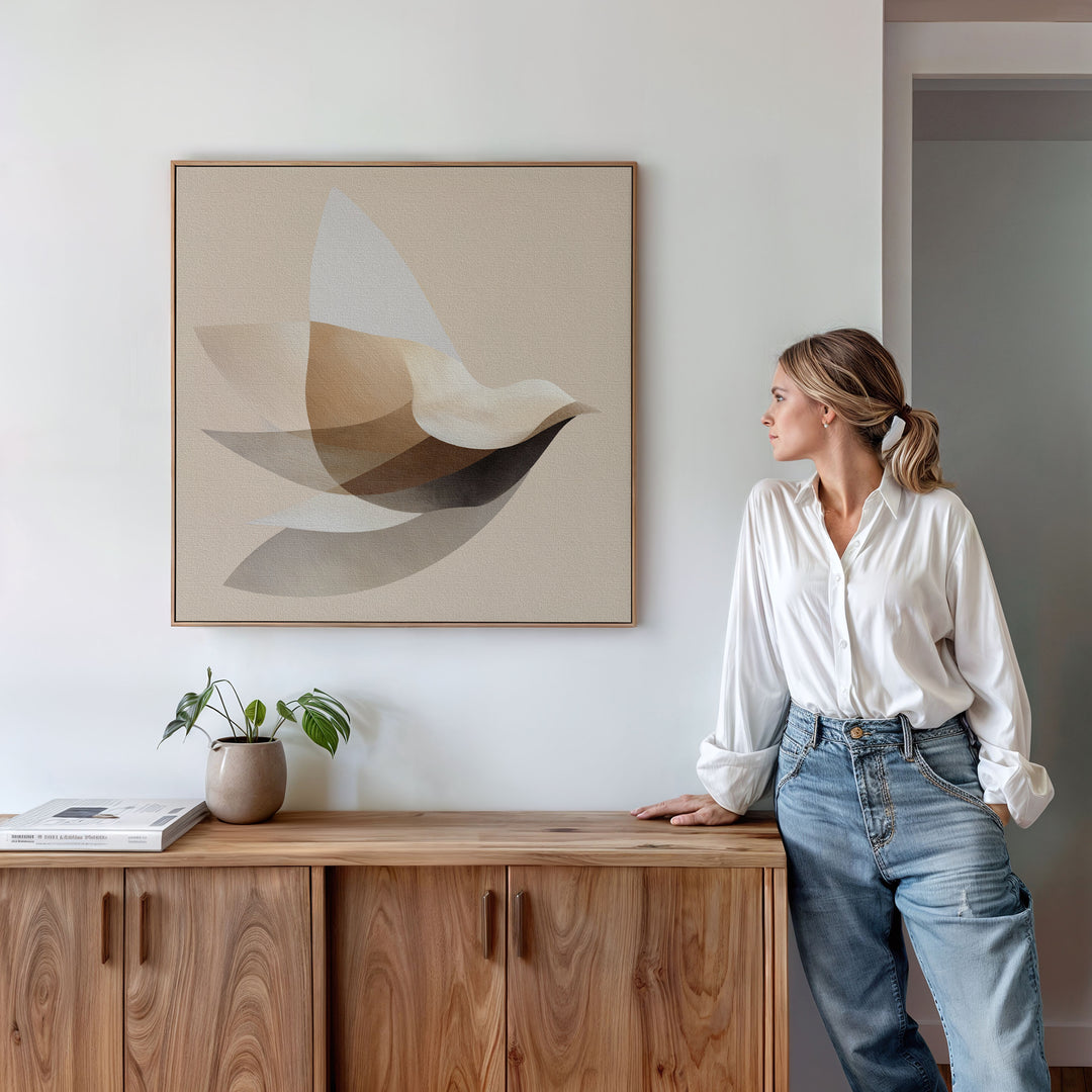 A woman in a white shirt and blue jeans leans against a wooden cabinet, admiring the Soaring Stillness Canvas Art. A small plant and books enhance the modern living room decor on the cabinet.
