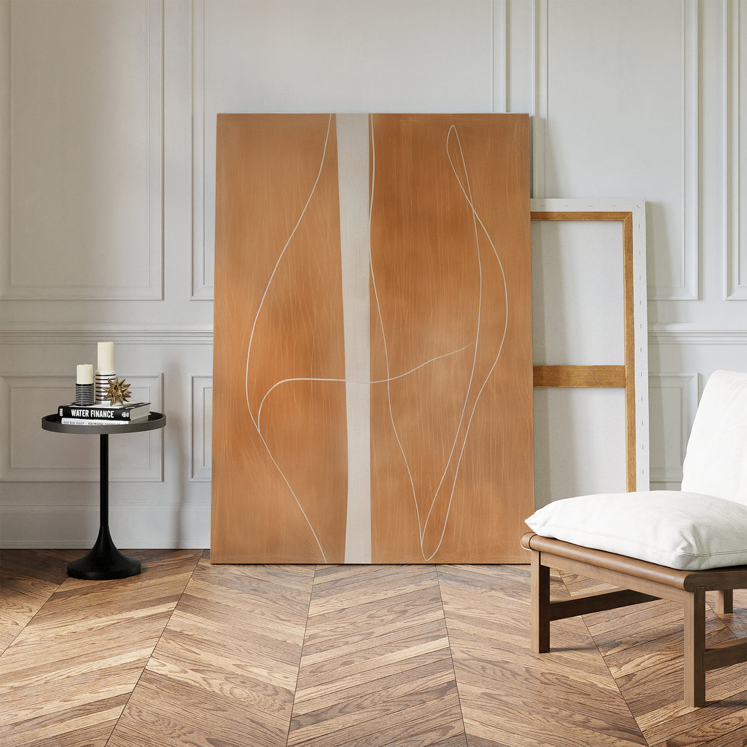The Soft Brown Flow Abstract Canvas Art, featuring curved white lines on a large wooden canvas, leans against a white paneled wall—ideal minimalist wall art for modern living rooms. Nearby are candles on a black table and a contemporary chair.