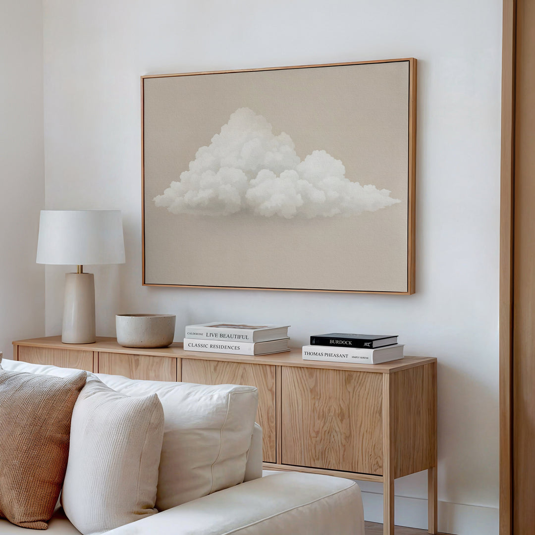 A modern, minimal living room with a wooden sideboard, white lamp, decorative bowl, and stacked books. The Soft Drift Canvas Art of fluffy clouds is framed above a cream sofa against a crisp white wall in neutral tones.
