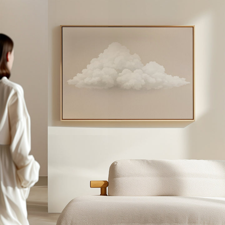 A person in white stands in a minimalist room, gazing at the Soft Drift Canvas Art—a fluffy white cloud on a beige wall above a light sofa—adding subtle charm and neutral tones to the space.