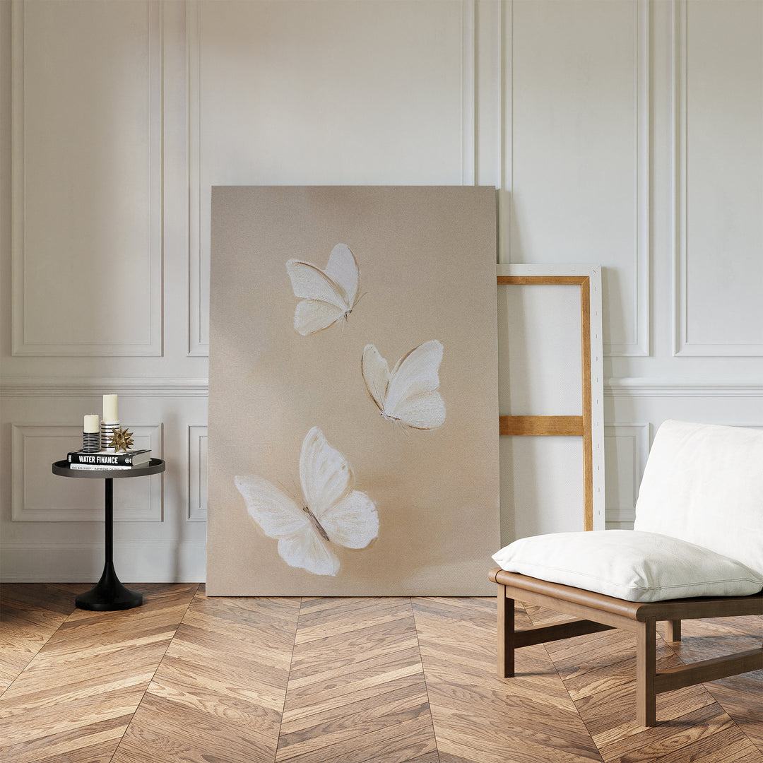 The Linen Wings Canvas Art, showcasing three white butterflies, leans against a white paneled wall. Beside it, a round black table with candles and books and a cushioned wood chair sit on a herringbone wood floor.