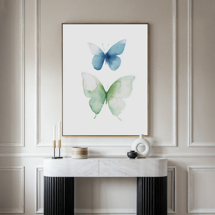 The Soft Flight Canvas Art, a minimalist piece with blue and green watercolor butterflies, is framed and displayed above a marble-topped Nordic Park console decorated with candles, books, and a sculptural white vase.