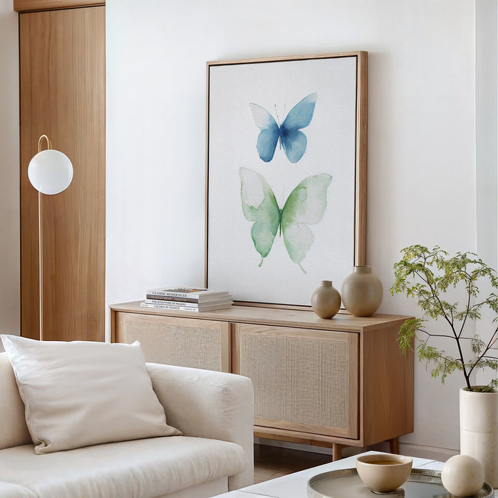 A modern living room with a light wood sideboard, neutral decor, and Soft Flight Canvas Art featuring blue and green watercolor butterflies. A white sofa and sleek table are in the foreground, with a potted plant and books on the sideboard.