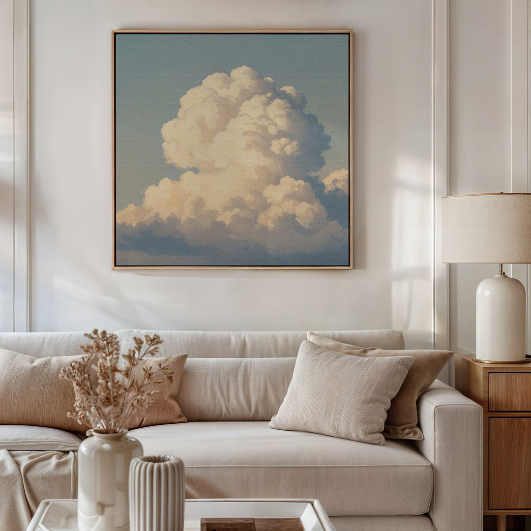 A cozy, modern living room with a beige sofa, neutral pillows, a dried flower arrangement on the coffee table, a table lamp, and the Soft Skies Rising Canvas Art—a large cloudscape print—displayed above the sofa.