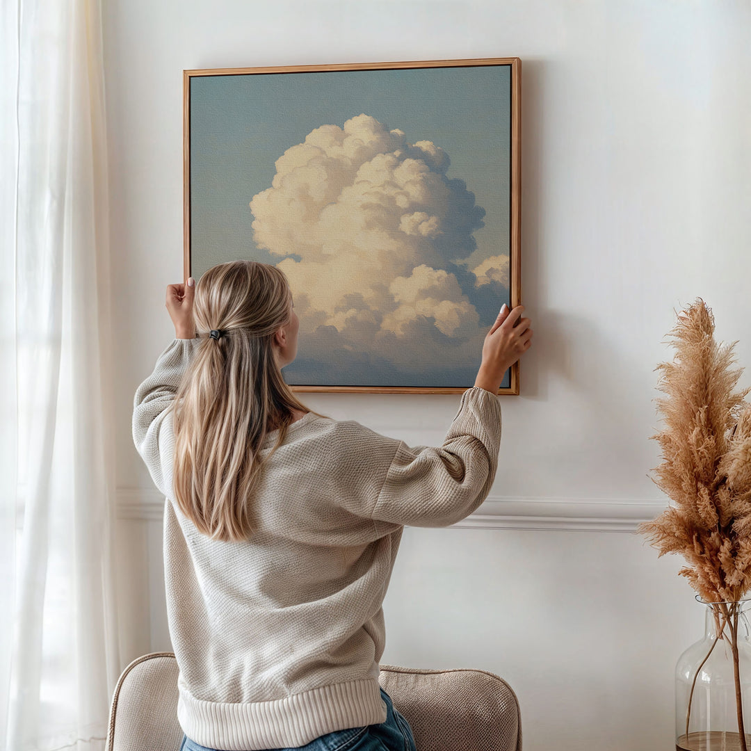 A woman with long blonde hair hangs the Soft Skies Rising Canvas Art on a white wall in a softly lit, neutral room with dried pampas grass, creating serene Nordic vibes.