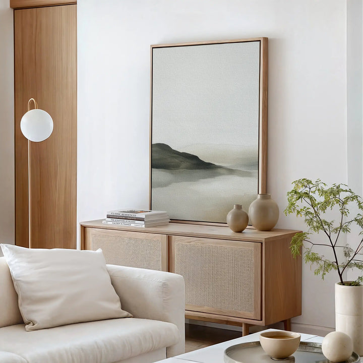 Minimalist living room with light wood furniture, a white sofa with cushions, a sideboard holding vases and books, minimalist decor, Soft Valley Canvas Art – Vertical on the wall, a spherical lamp, and a small green plant.