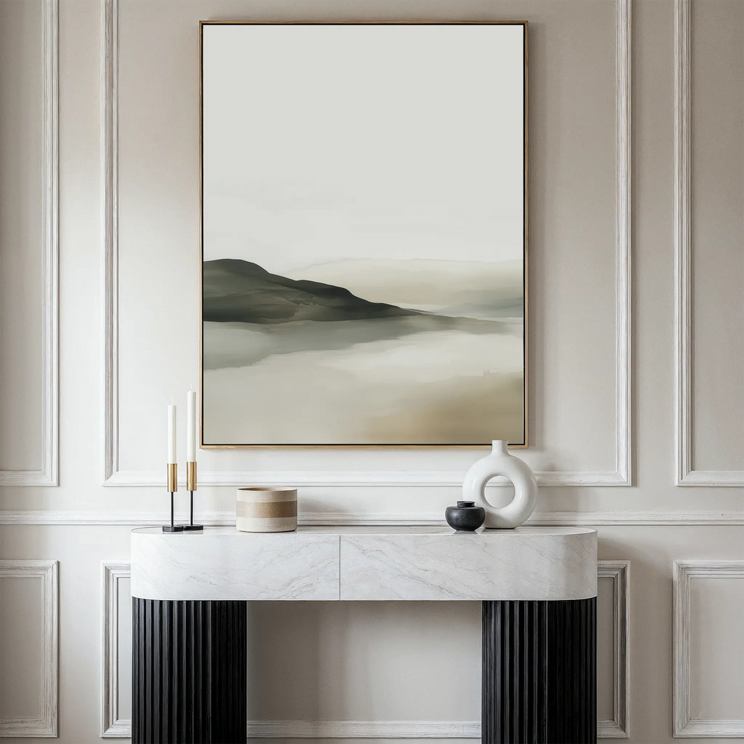 The Soft Valley Canvas Art – Vertical, featuring a minimalist misty mountain landscape, hangs above a modern marble-top console table decorated with two candles, a beige container, a black vase, and a white ceramic sculpture.