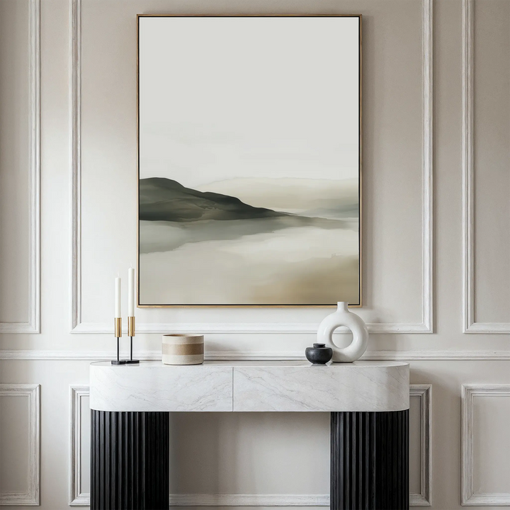 The Soft Valley Canvas Art – Vertical, featuring a minimalist misty mountain landscape, hangs above a modern marble-top console table decorated with two candles, a beige container, a black vase, and a white ceramic sculpture.