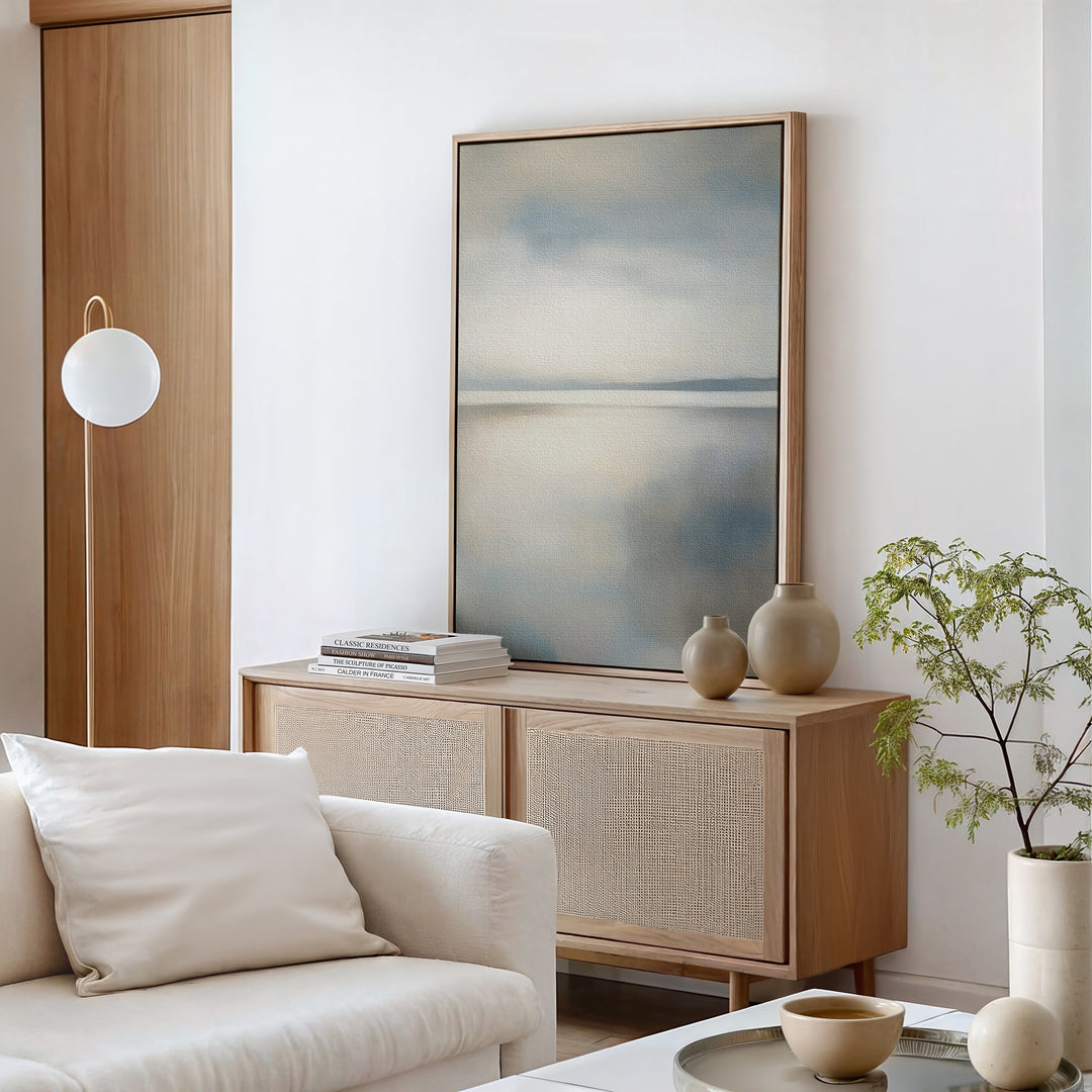 A minimalist living room with a light wood sideboard, neutral vases, books, a potted plant, and a white sofa with cushion—all in soft tones—features the Still Horizon Canvas Art, adding serene shoreline style to the space.