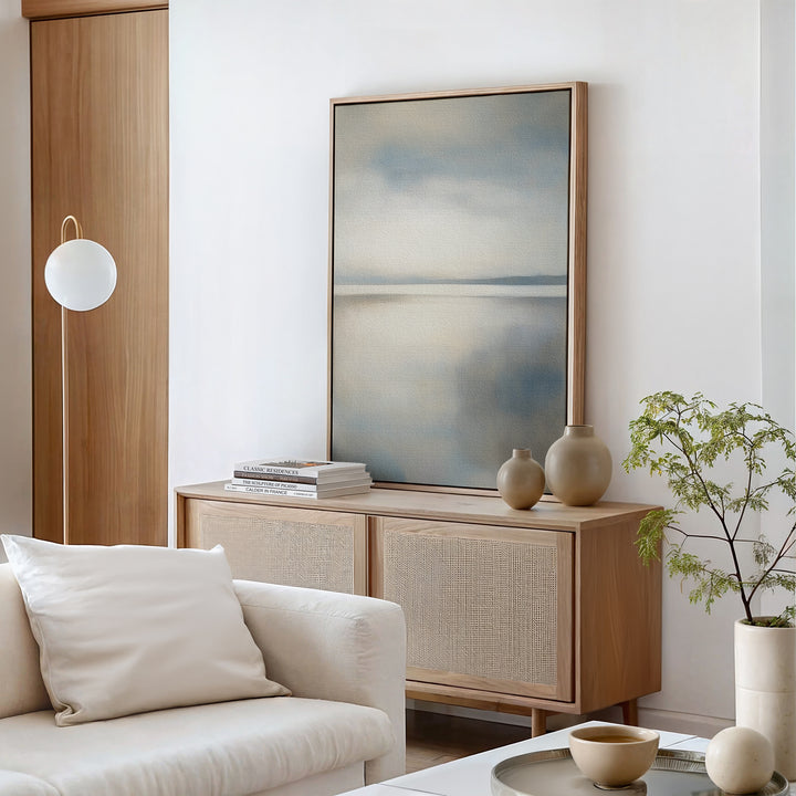 A minimalist living room with a light wood sideboard, neutral vases, books, a potted plant, and a white sofa with cushion—all in soft tones—features the Still Horizon Canvas Art, adding serene shoreline style to the space.