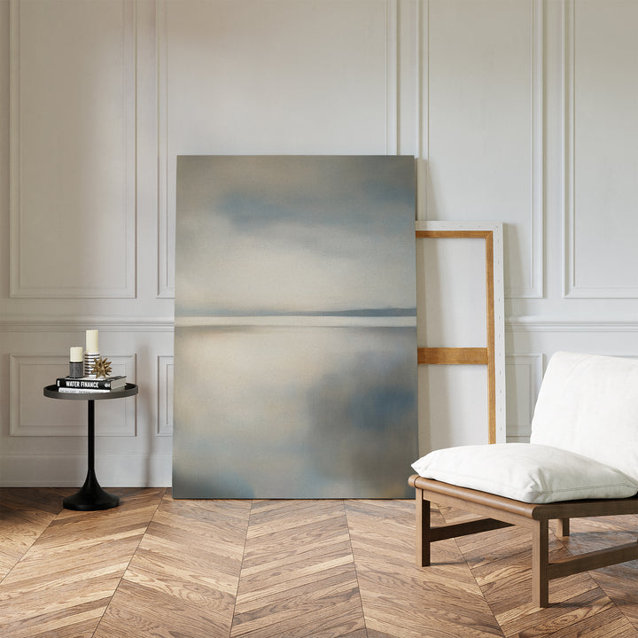 A minimalist room with white paneled walls, herringbone wood floor, a low cushioned chair, and a circular side table with candles and books features two large canvases. The Still Horizon Canvas Art adds calm, minimalist wall decor to the space.