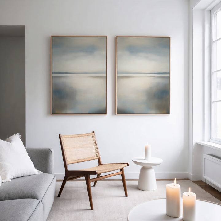 A minimalist living room with a light gray sofa, woven wooden chair, small white side table with a candle, three lit candles on a white coffee table, and the Still Horizon Set of 2 as serene minimalist wall art above.