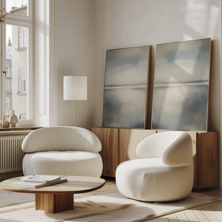 A modern living room with two rounded white armchairs, a low wooden coffee table, a minimalist floor lamp, and the Still Horizon Set of 2 serene shoreline art leaning on a wooden sideboard. Soft natural light fills the space.