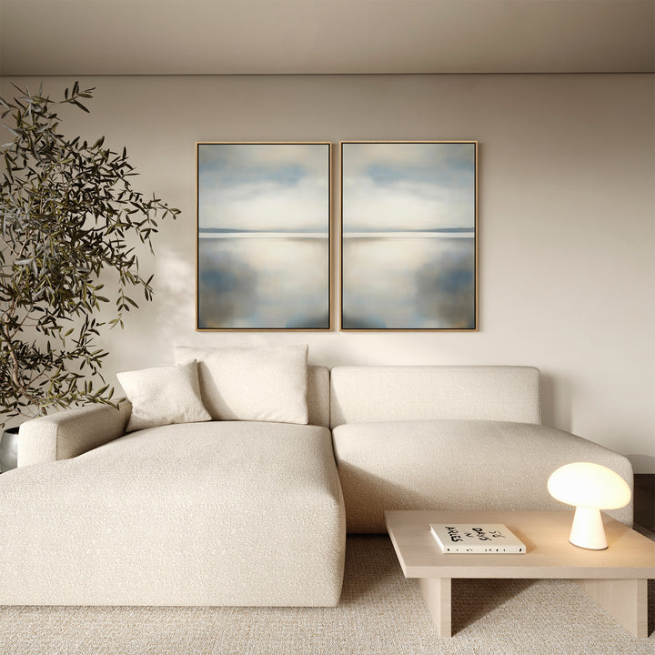 A minimalist living room features a cream sectional, light wood coffee table, tall potted plant, and the Still Horizon Set of 2 wall art in abstract blue and beige, evoking the calm of a serene shoreline.