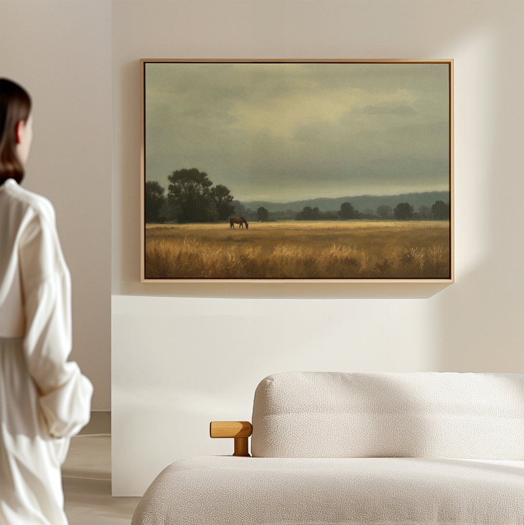 A woman in a white outfit stands in a minimalist room, admiring the Still Morning Canvas Art—a framed landscape of a horse grazing in a golden field under cloudy skies. A light sofa completes the serene Nordic Park-inspired setting.