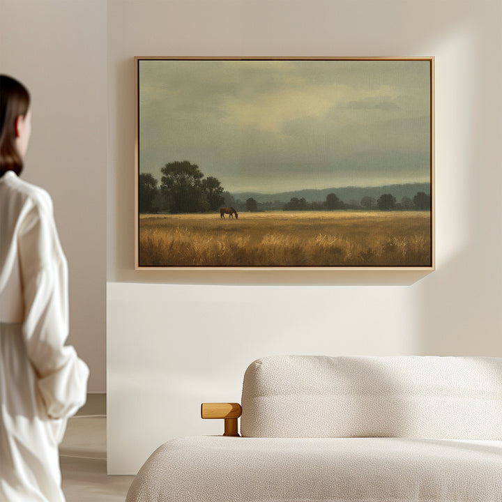 A woman in a white outfit stands in a minimalist room, admiring the Still Morning Canvas Art—a framed landscape of a horse grazing in a golden field under cloudy skies. A light sofa completes the serene Nordic Park-inspired setting.