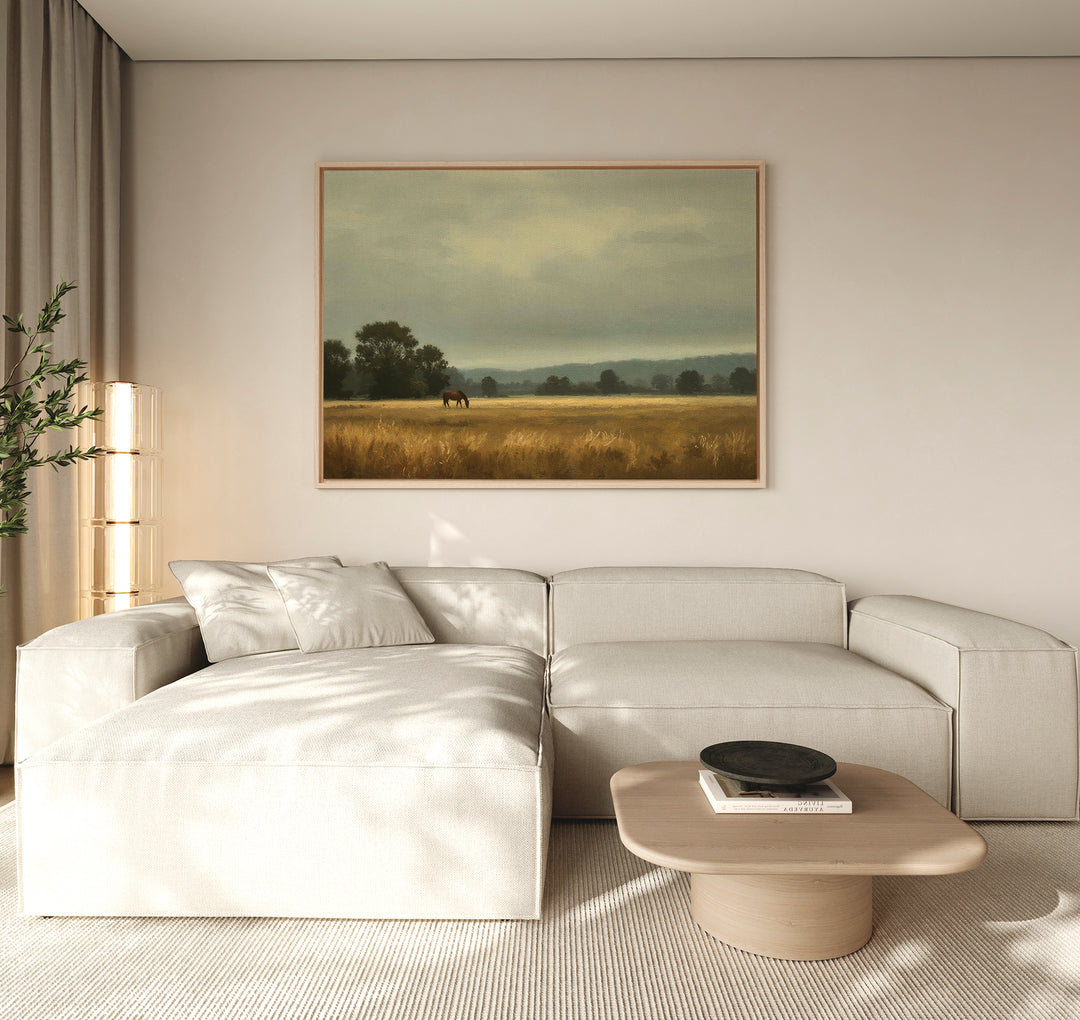 A modern living room features a beige sectional sofa, a light wood coffee table, and the Still Morning Canvas Art depicting a field with trees and a horse. Tall curtains let in natural light, enhancing the serene atmosphere.
