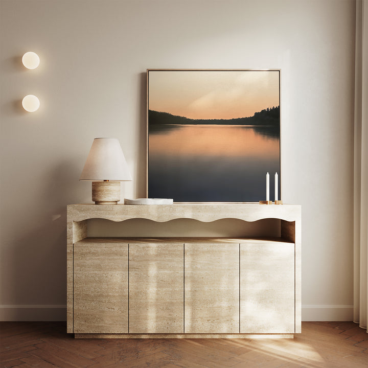 A minimalist console table displays a lamp, two candles, and the Still Waters Canvas Art depicting a tranquil sunset lake. Two round wall lights are on the left, with soft natural light on the right—perfect living room decor.