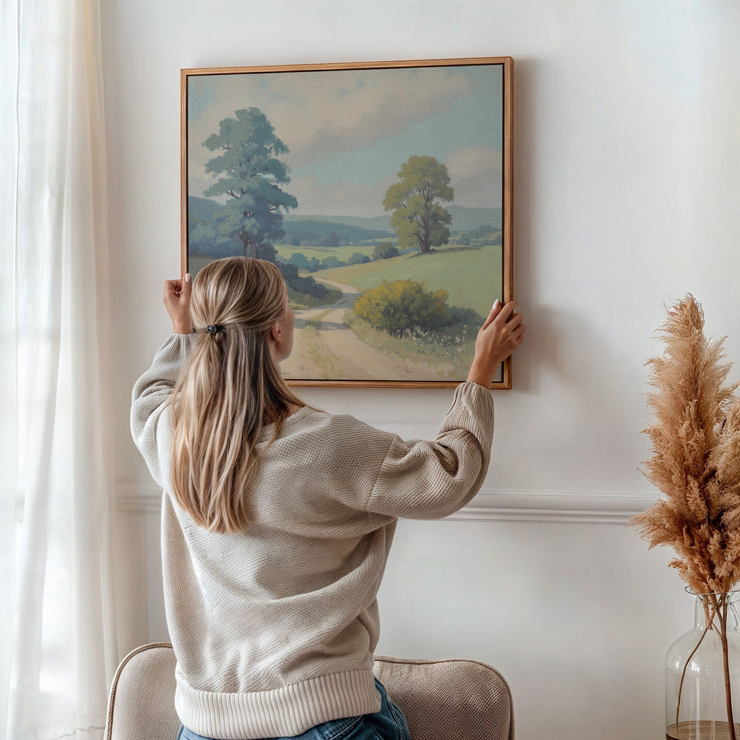 A woman with long blonde hair in a ponytail hangs the Summer Dale Canvas Art, a framed countryside landscape, on a white wall behind a beige chair. Dried pampas grass in a vase nearby adds rustic wall art charm to the space.