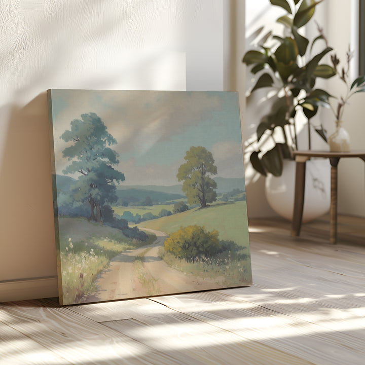 The Summer Dale Canvas Art rests against a sunlit wall, showcasing a winding path, green fields, and tall trees. Nearby, a plant and small table sit on the light wooden floor as soft, natural light fills the room.