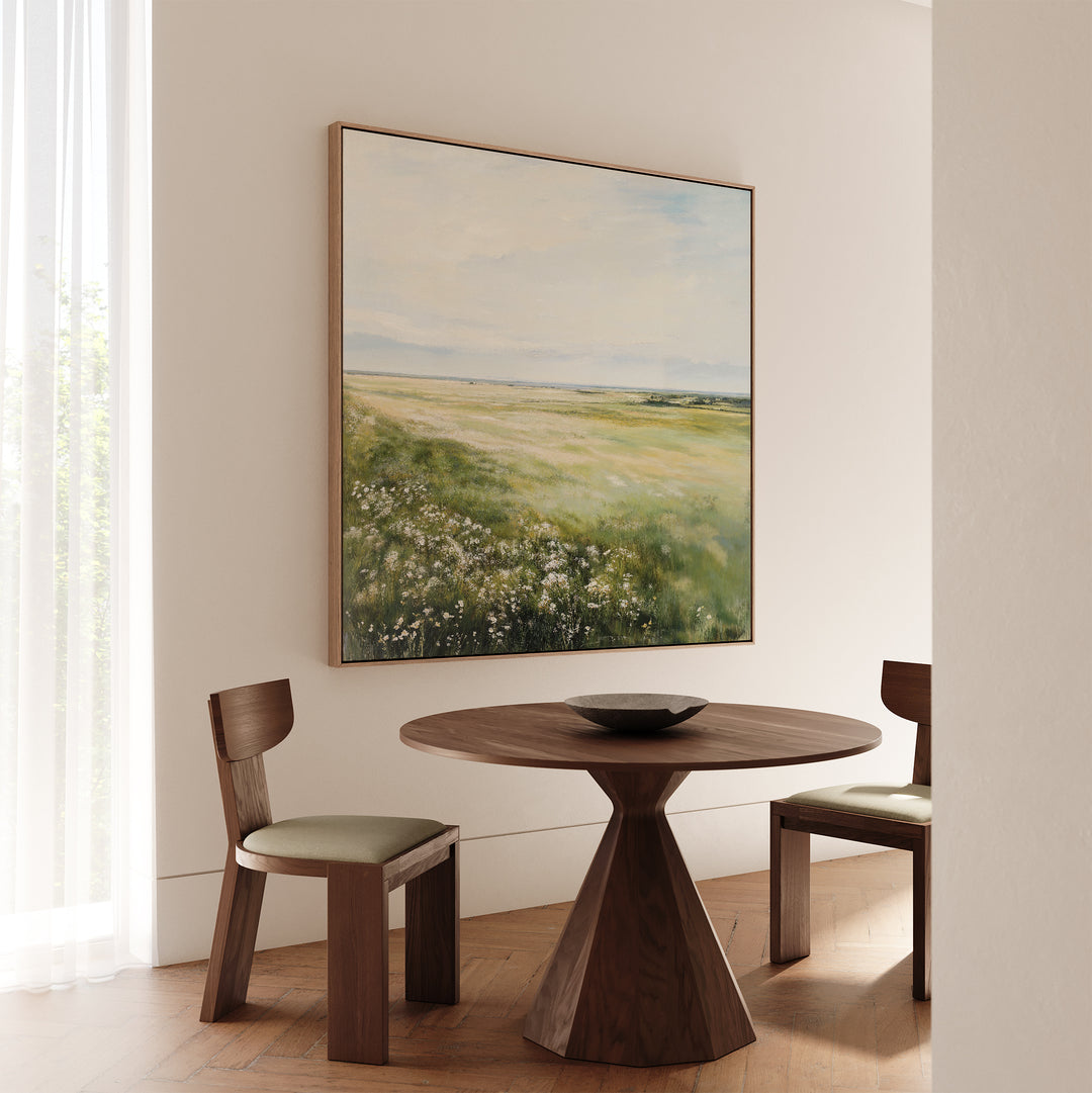 A round wooden table with two matching chairs sits near a window with sheer curtains, adding warmth to the living room. Above the table, the Summer Meadow Canvas Art brings a touch of nature’s beauty into the space.