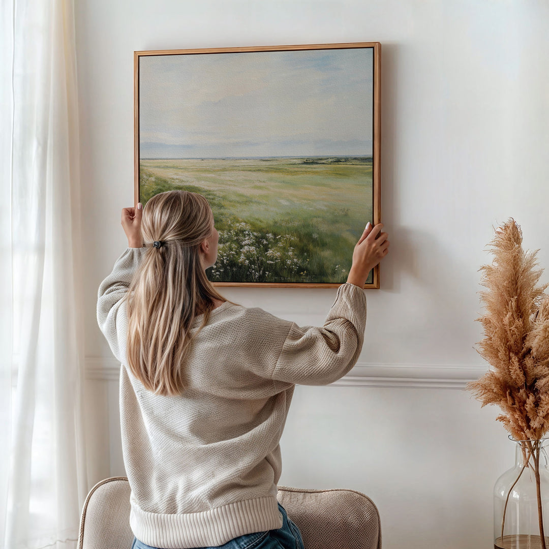 A woman with long blonde hair hangs the Summer Meadow Canvas Art, featuring a grassy field and sky, on a white wall in a cozy, light-filled room with beige decor and a vase of pampas grass, celebrating nature’s beauty.