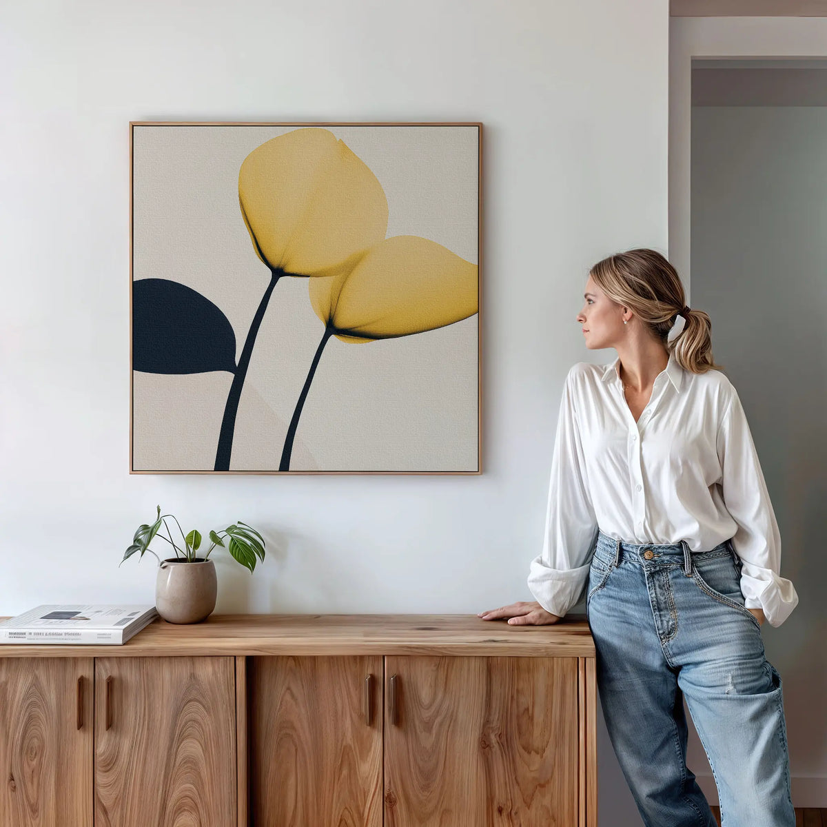 A woman in a white blouse and jeans stands by a wooden cabinet in a modern living room, admiring the Sunny Flora Canvas Art with yellow blooms on the wall. A potted plant and books sit atop the cabinet.
