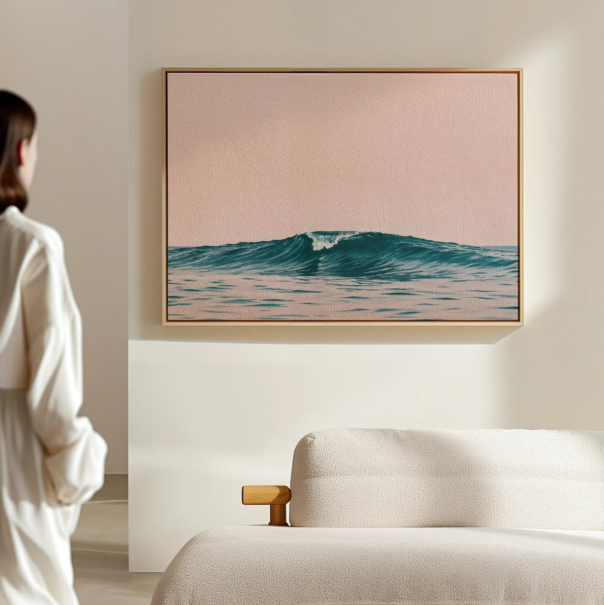 A person stands in a minimal, light-filled room beside a cream-colored sofa, admiring the Sunset Wave Canvas Art—a framed ocean wave beneath a soft pink sky—serving as beach-inspired wall decor.