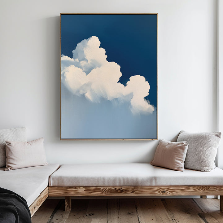 A modern living room with light wood furniture and neutral cushions, highlighted by calming decor and The Cumulus Abstract Canvas Art—a large framed painting of fluffy white clouds on a deep blue sky—adding minimalist style to the white wall.