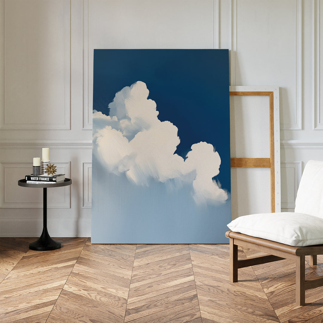 The Cumulus Abstract Canvas Art, featuring fluffy white clouds against a blue sky, leans against a white wall and adds a calming touch to a room with herringbone floors, a cushioned chair, an empty frame, and a table with candles and books.