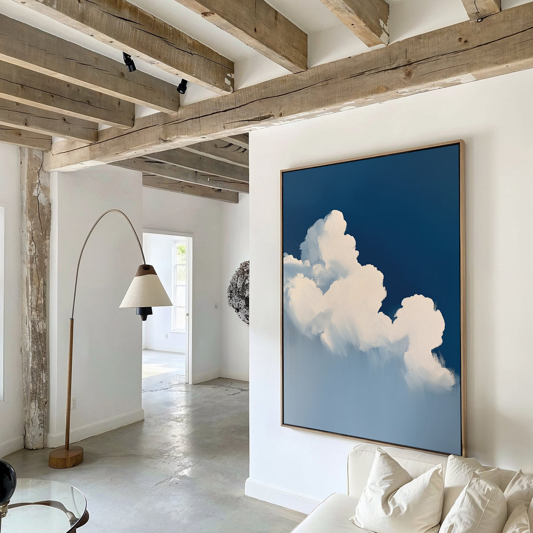 A modern living room with exposed wooden beams, white walls, a floor lamp, and calming decor featuring The Cumulus Abstract Canvas Art—large wall art of fluffy white clouds against a deep blue sky—hanging on the wall.
