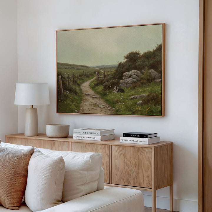 A cozy living room features a wood sideboard with stacked books, a lamp, and ceramics. Above it hangs The Watchful Path Canvas Art, depicting a grassy rural path with rocks and a dog, bringing rustic charm to the space.