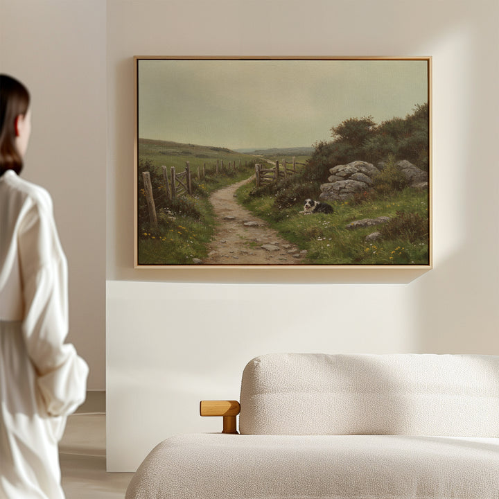 A woman in a white robe stands in a minimalist room, admiring The Watchful Path Canvas Art—a painting of a path with a dog on grassy fields by rocks and a fence. A beige sofa enhances the rustic living room decor.