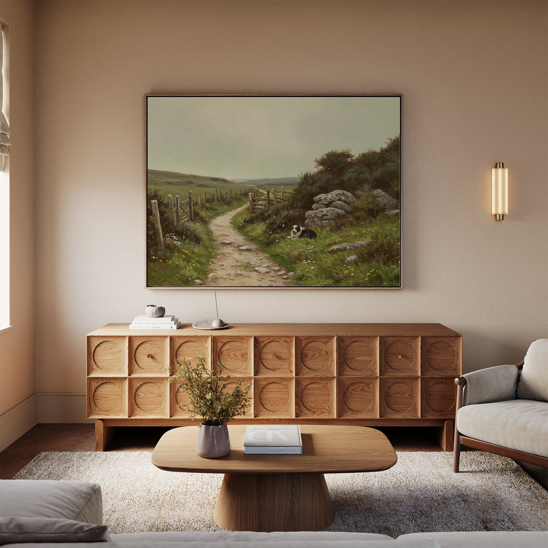 A cozy living room with a light beige sofa, wooden coffee table, textured wooden sideboard, and The Watchful Path Canvas Art—a landscape painting—above the sideboard. Small plants and books accent the tables.