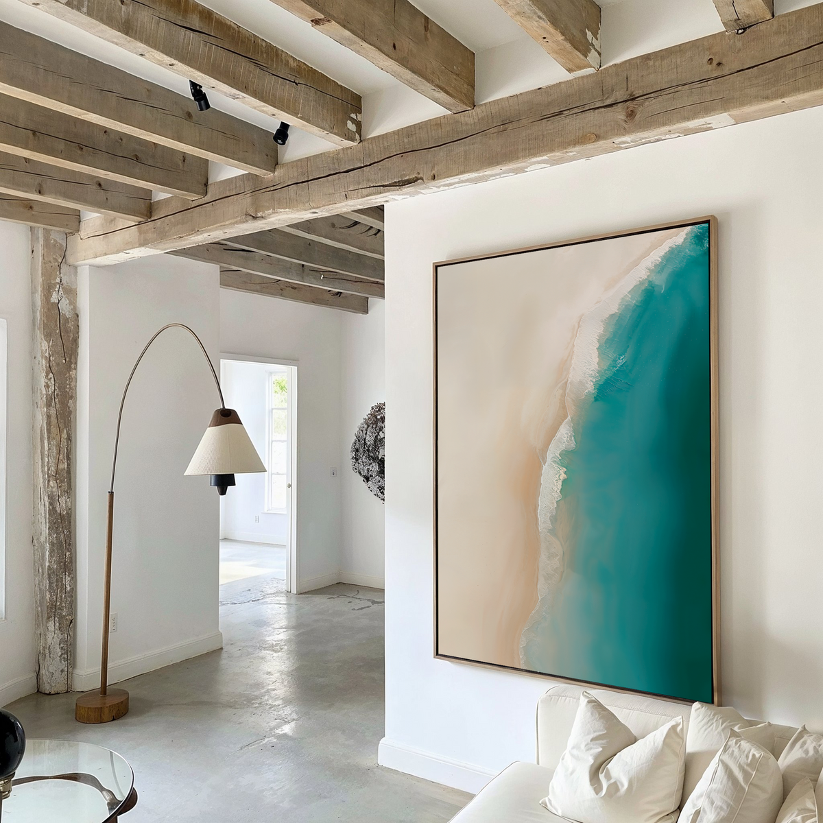 A modern living room with exposed wooden beams, white walls, a curved floor lamp, and The Beach Abstract Canvas Art—a large framed piece showing an aerial view of turquoise sea meeting sandy shore.