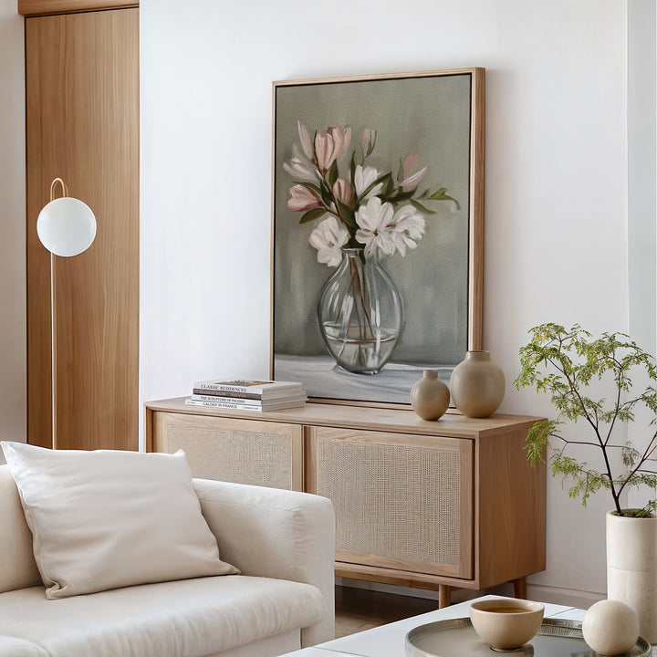 A minimalist living room showcases a wooden sideboard with vases, stacked books, and the Vase & Grace Canvas Art—white and blush flowers in a glass vase. A white sofa with cushion and a potted plant complete the serene decor.